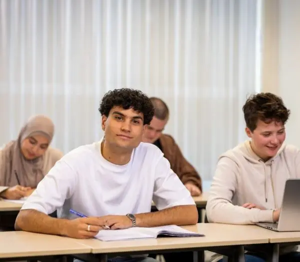 Studenten in klaslokaal. Jongen vooraan kijkt in de camera