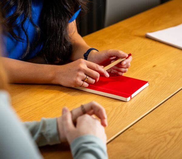 Een rood boekje ligt op tafel, de handen van een student zijn zichtbaar met een pen in de hand
